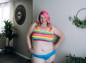 Chrissy with neon rainbow and pink hair, wearing a neon rainbow crop top and turquoise underwear